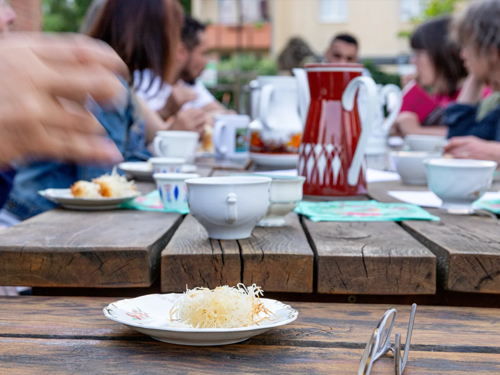 Kaffeetafel im Freien mit Tassen, Kannen und Kuchen, im Hintergrund Gäste